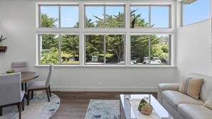 A room with a large window, a dining table, chairs, a sofa, and a coffee table. - Miramar Oasis! Ocean View, Hot Tub, Steps2Beach, Roof Deck, Fire Pit, Coastal Trail (El Granada)
