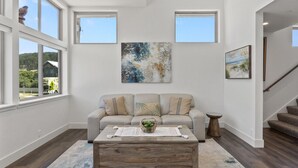 A modern living room with a beige sofa, a wooden coffee table, and artwork on the walls. - Miramar Oasis! Ocean View, Hot Tub, Steps2Beach, Roof Deck, Fire Pit, Coastal Trail (El Granada)