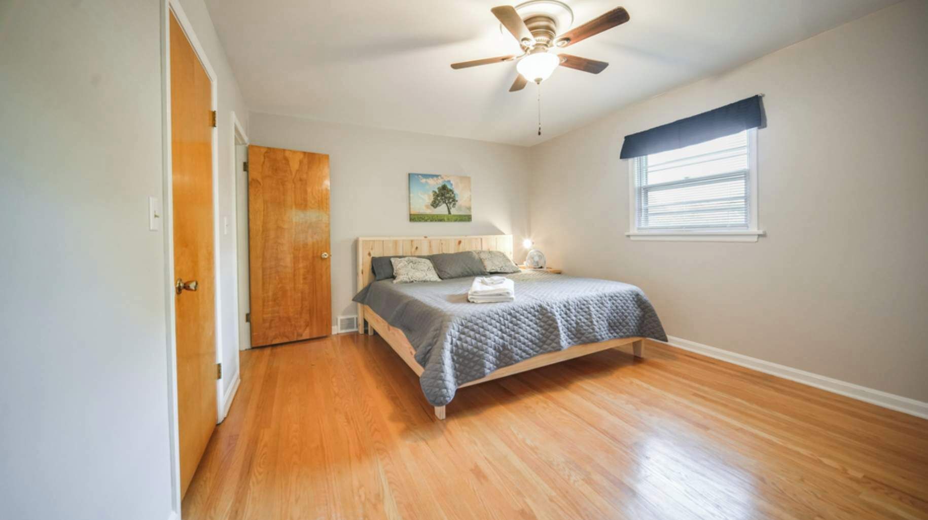 Un dormitorio con cama, piso de madera, ventilador de techo, ventana con persianas y puerta.
