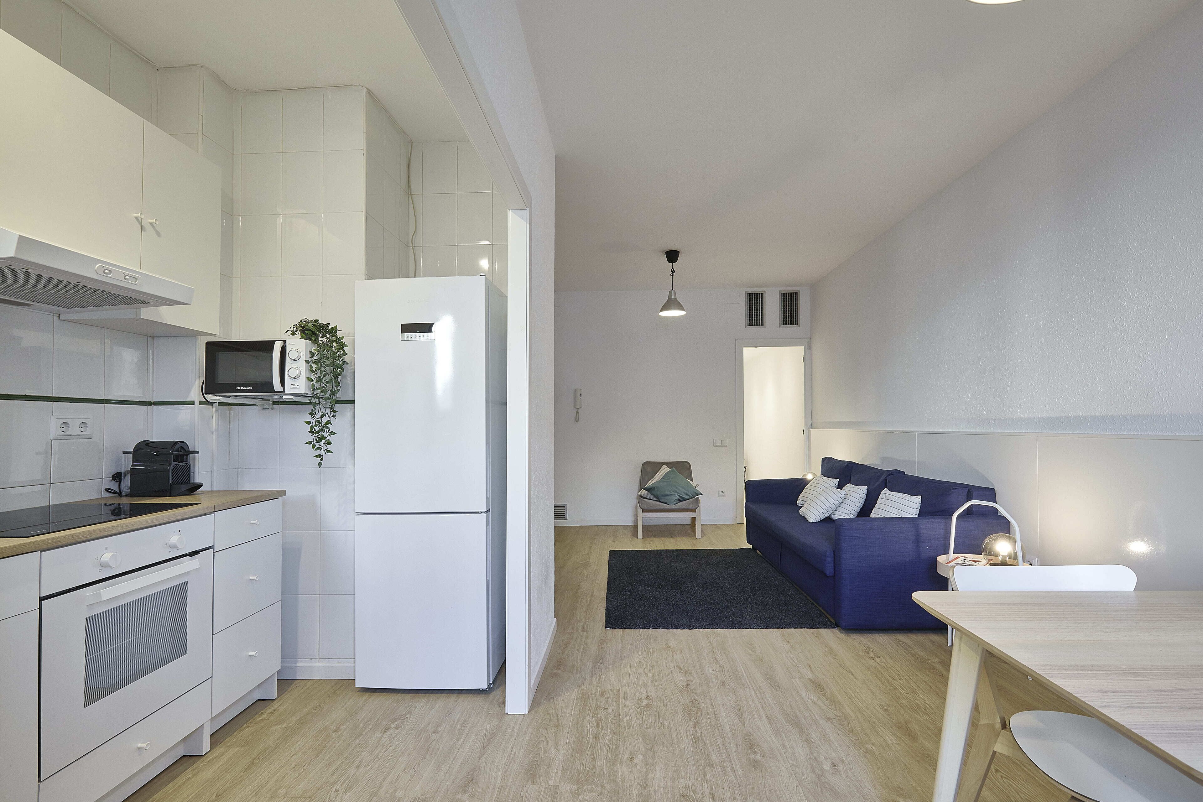 Eine moderne Küche mit weißen Schränken, einer Mikrowelle und einem Kühlschrank. Es gibt einen Essbereich mit Tisch und Stühlen, und im Hintergrund steht ein blaues Sofa.