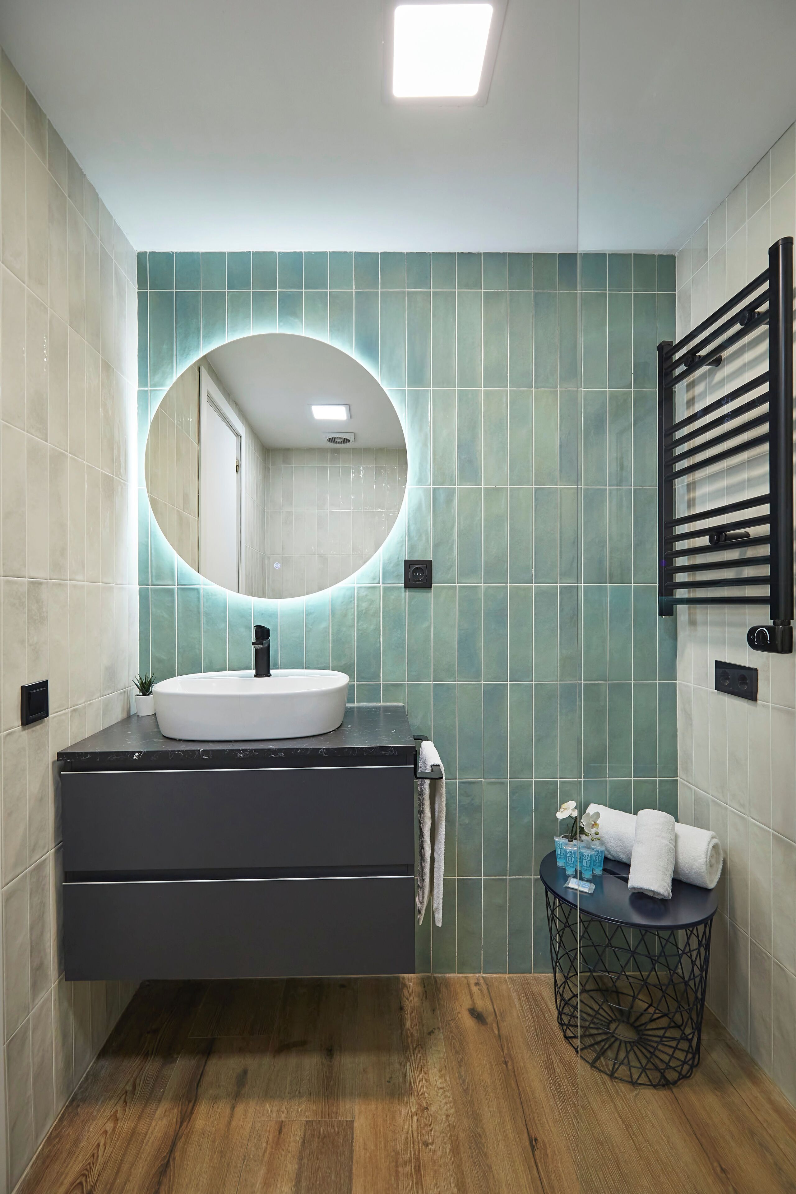 A bathroom with a round mirror, a white sink, a towel rack, and a small round table with a towel and a plant.