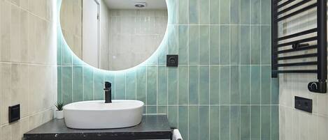 A bathroom with a round mirror, a white sink, a towel rack, and a small round table with a towel and a plant.