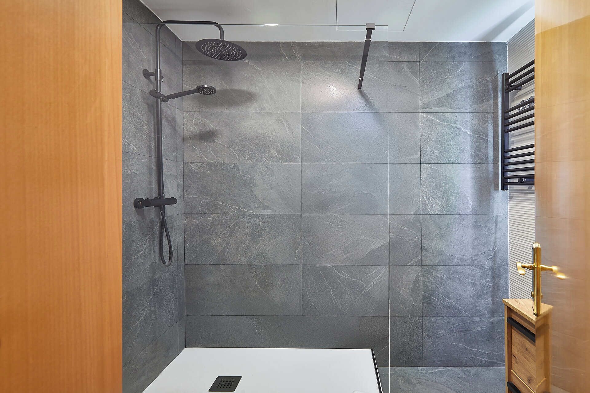 A shower with a handheld showerhead and a wall-mounted head, tiled with grey stone.