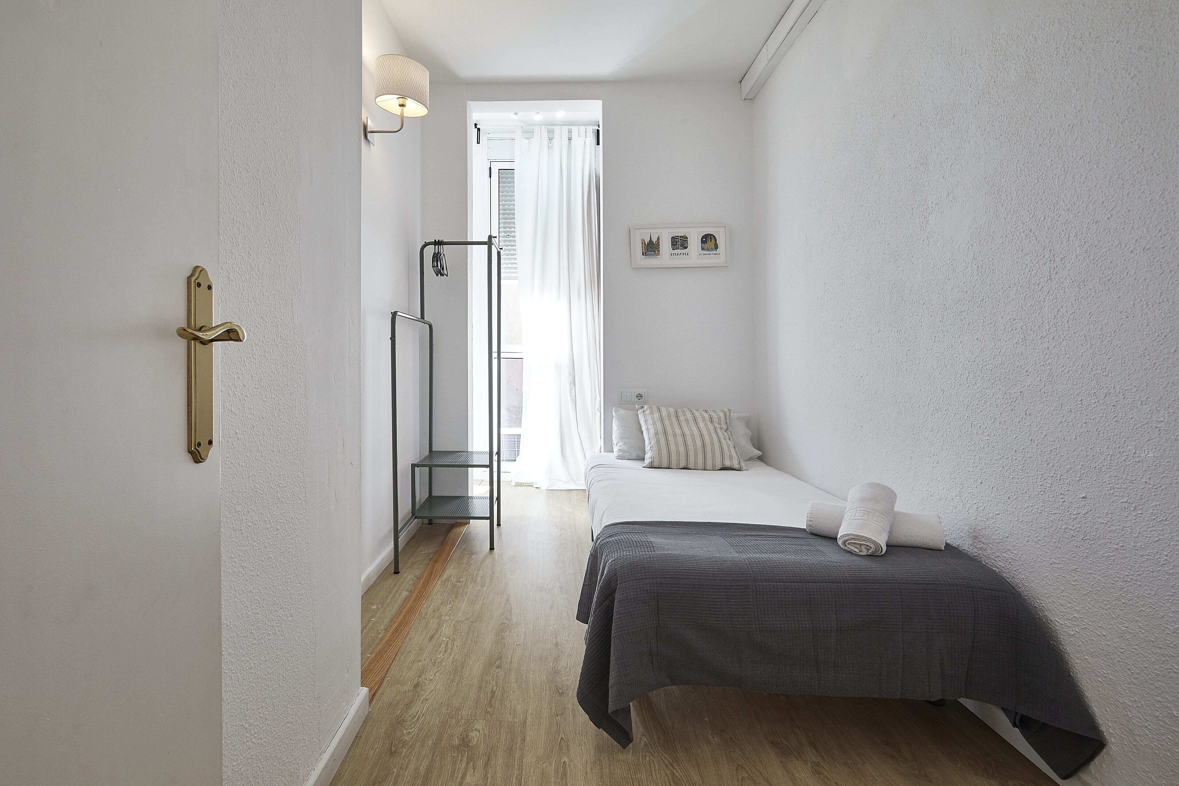 Een slaapkamer met een bed, nachtkastje, hanglamp, raam met gordijnen en een deur met handgreep.