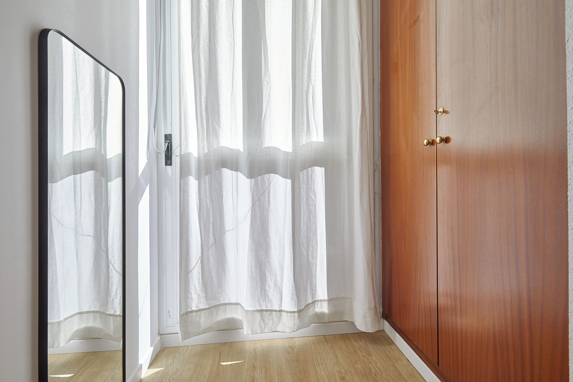 Ein Zimmer mit Holzschrank, durchsichtigen Vorhängen und einem Spiegel, der das Innere widerspiegelt.