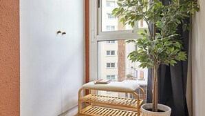 Ein Eckzimmer mit weißer Wand, einem Fenster mit Blick auf Gebäude, einer hölzernen Bank mit Kissen und einer Topfpflanze.