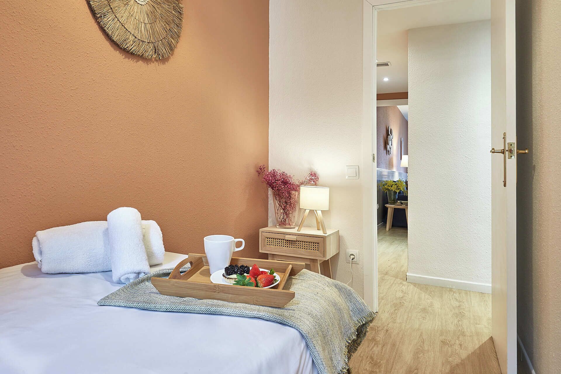 A hotel room with a bed, a tray with strawberries, a cup, and a nightstand with a lamp and flowers.