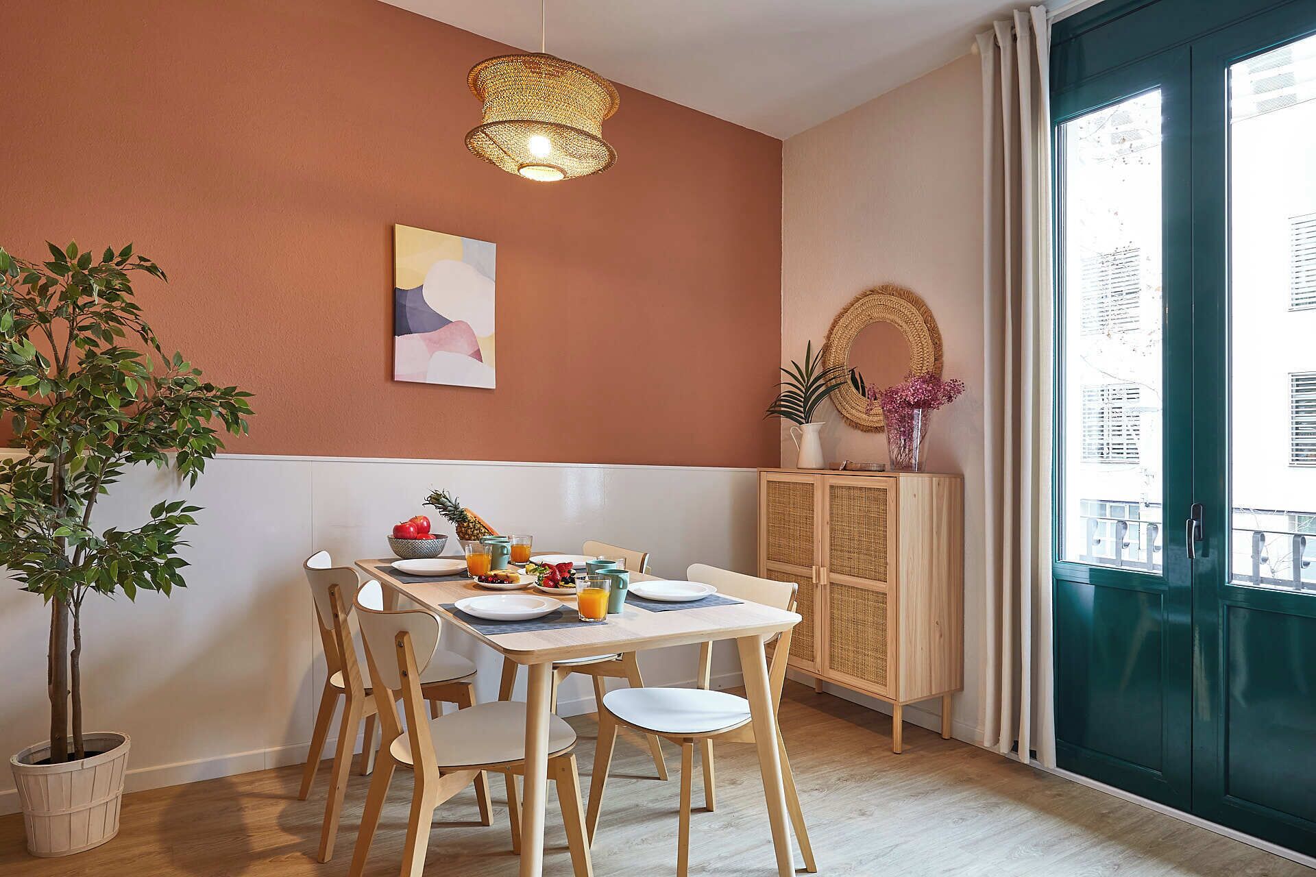 A dining area with a table set for four, a hanging light fixture, a painting on the wall, and a view of the outside through a large window.