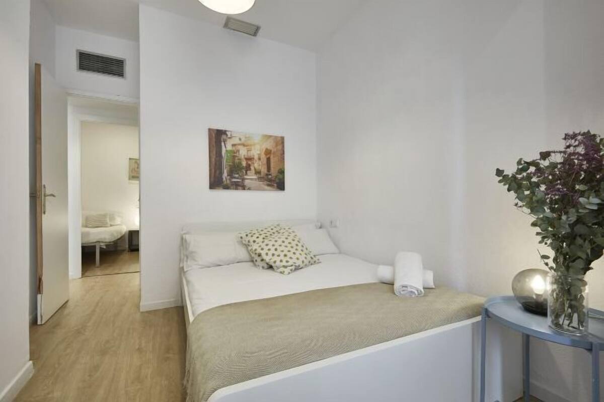 A bedroom with a bed, a bedside table with a vase, and a painting on the wall.