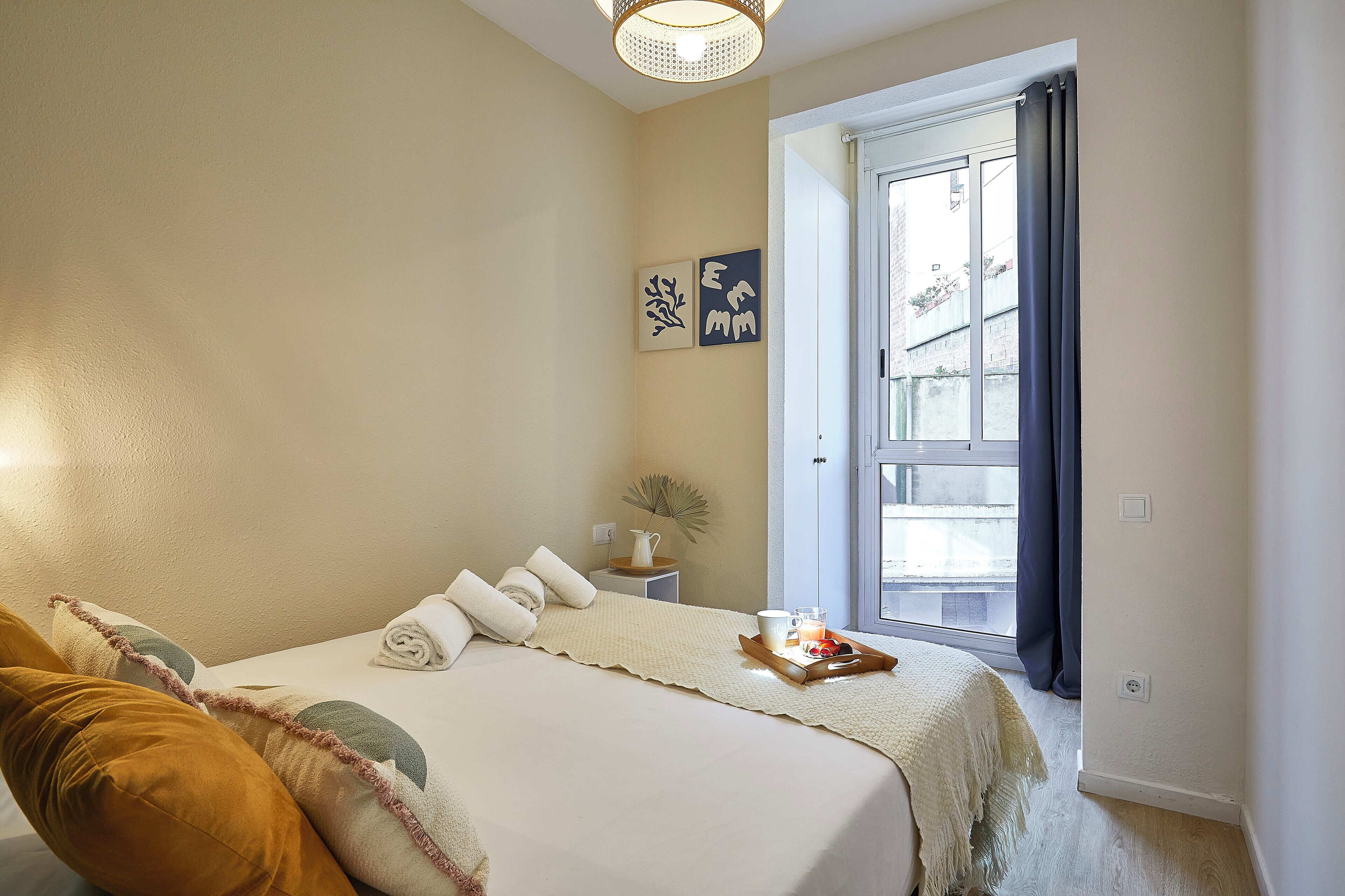 A bedroom with a bed, pillows, a bedside table with a lamp, a window with curtains, and artwork on the wall.