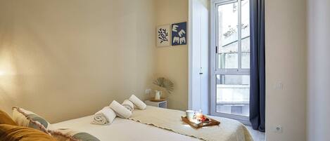 A bedroom with a bed, pillows, a bedside table with a lamp, a window with curtains, and artwork on the wall.