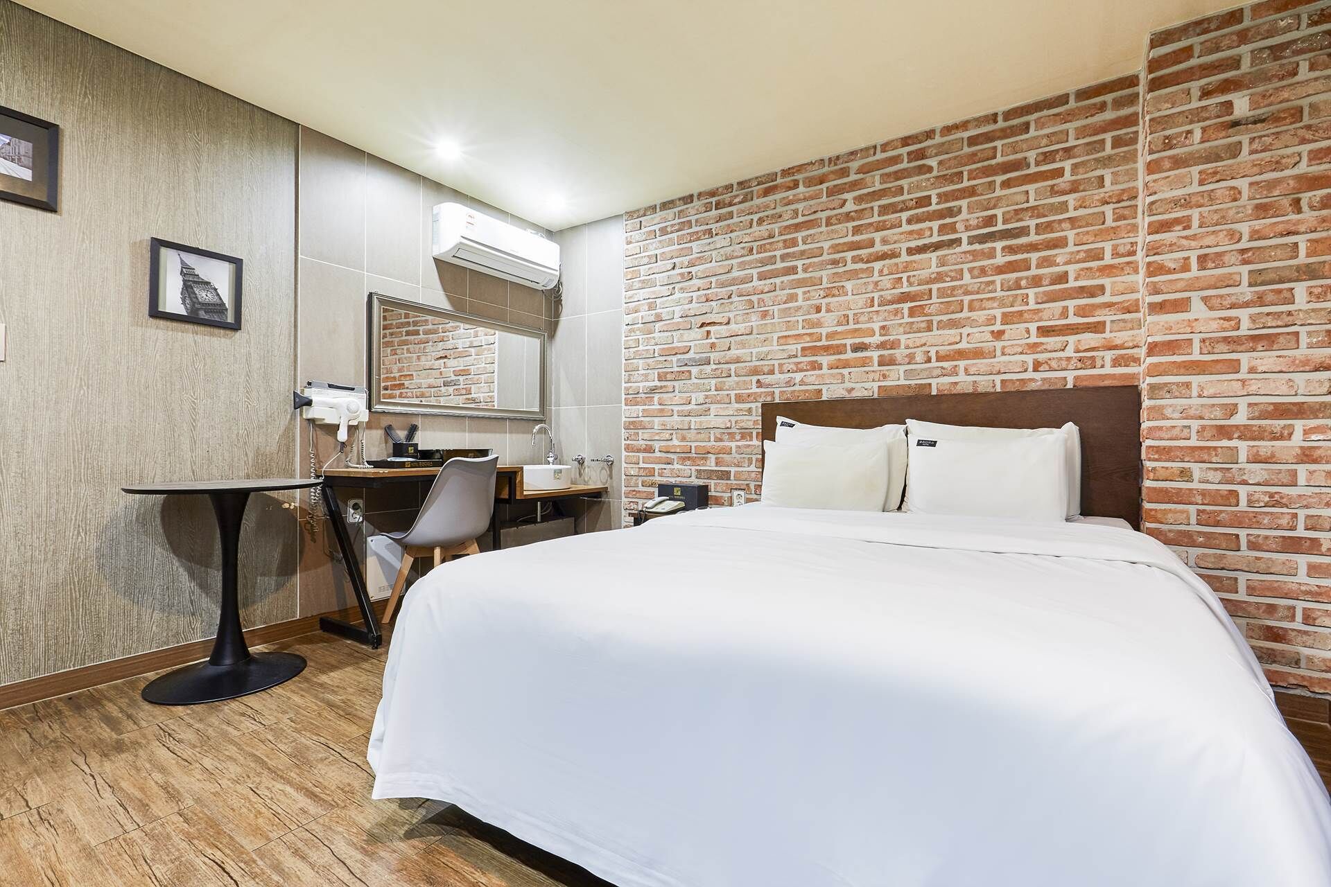 A hotel room with a brick wall, a bed with white bedding, a desk with a chair, and a mirror.