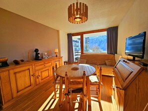 Una habitación acogedora con una mesa de madera, sillas, un sofá, un televisor y vistas a montañas a través de amplias ventanas.