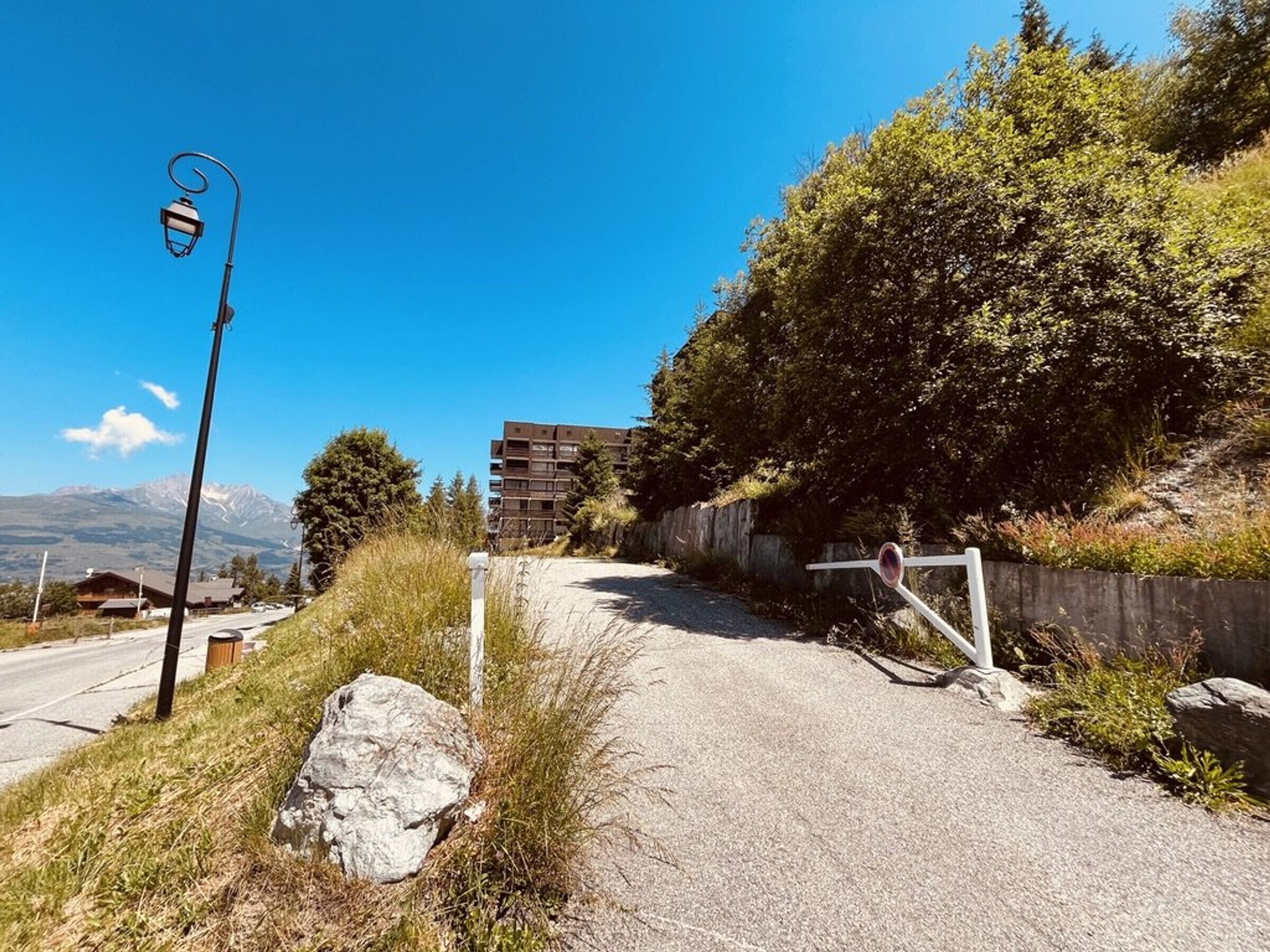 Ein Kiesweg führt zu einem Gebäude mit einem Metallgitter, umgeben von viel Grün und blauem Himmel.