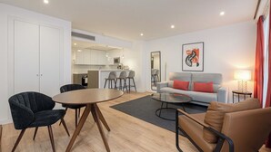 Un salon moderne comprenant un canapé, des fauteuils, une table basse et une table à manger. On aperçoit à l’arrière-plan un coin cuisine avec des tabourets de bar.