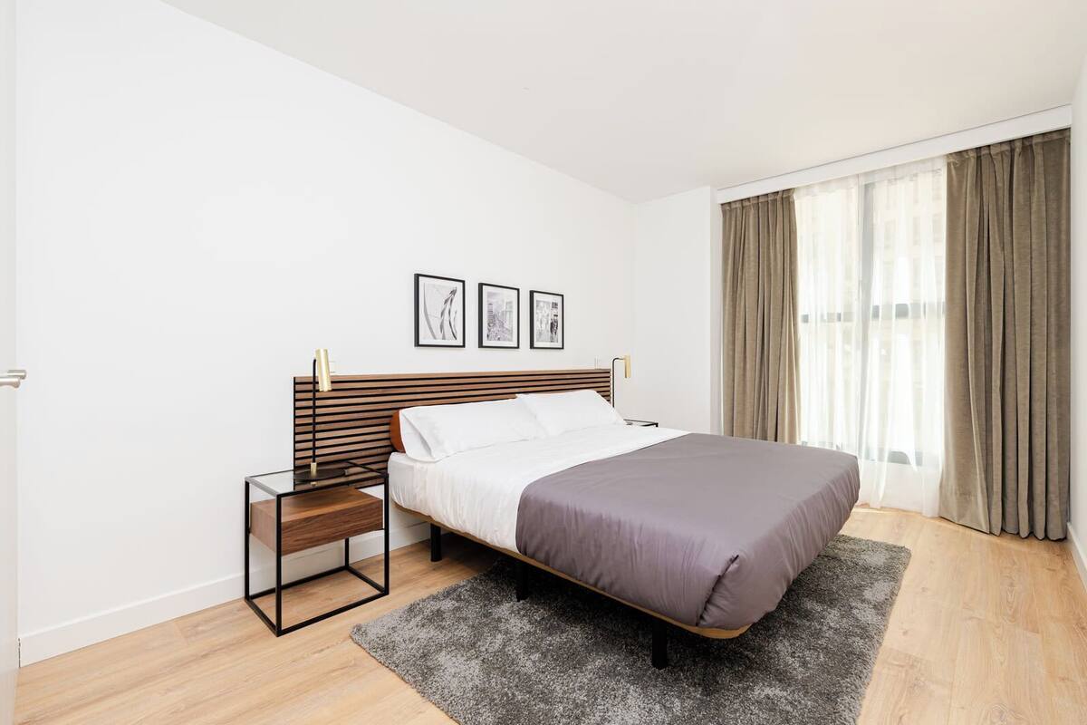 A bedroom with a bed, a nightstand, a window with curtains, and three framed pictures on the wall.
