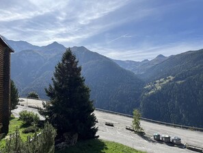 A mountain road with a wooden building on the left, a tall evergreen tree in the foreground, and a view of distant mountains under a partly cloudy sky.