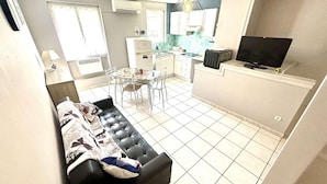 Una sala de estar con un sofá negro, un comedor con mesa y sillas, una cocina con armarios blancos y un televisor sobre un mueble.