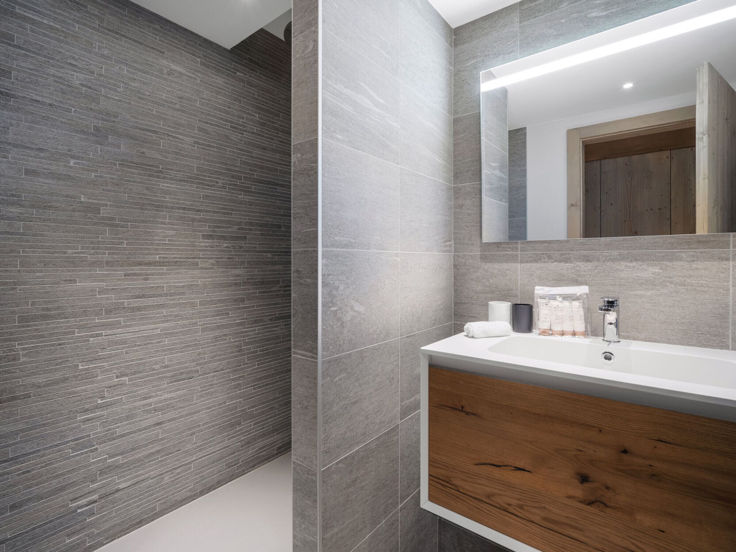 A modern bathroom with a grey brick wall, a wooden vanity, and a large mirror above the sink.