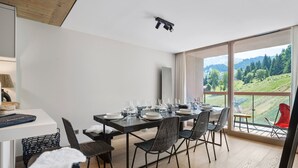Comedor con mesa dispuesta para seis personas, cuatro sillas y vista a un paisaje verde a través de amplias ventanas.