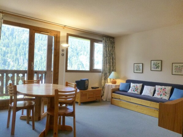 A cozy room with a wooden dining table, chairs, a sofa, and a small cabinet. Large windows offer a view of trees. - Charming 1-bedroom flat for 5 in Peisey Village center (Peisey-Vallandry)