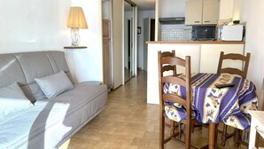 A living room with a grey sofa, a table with chairs, a lamp, and a kitchen area with cabinets and appliances.