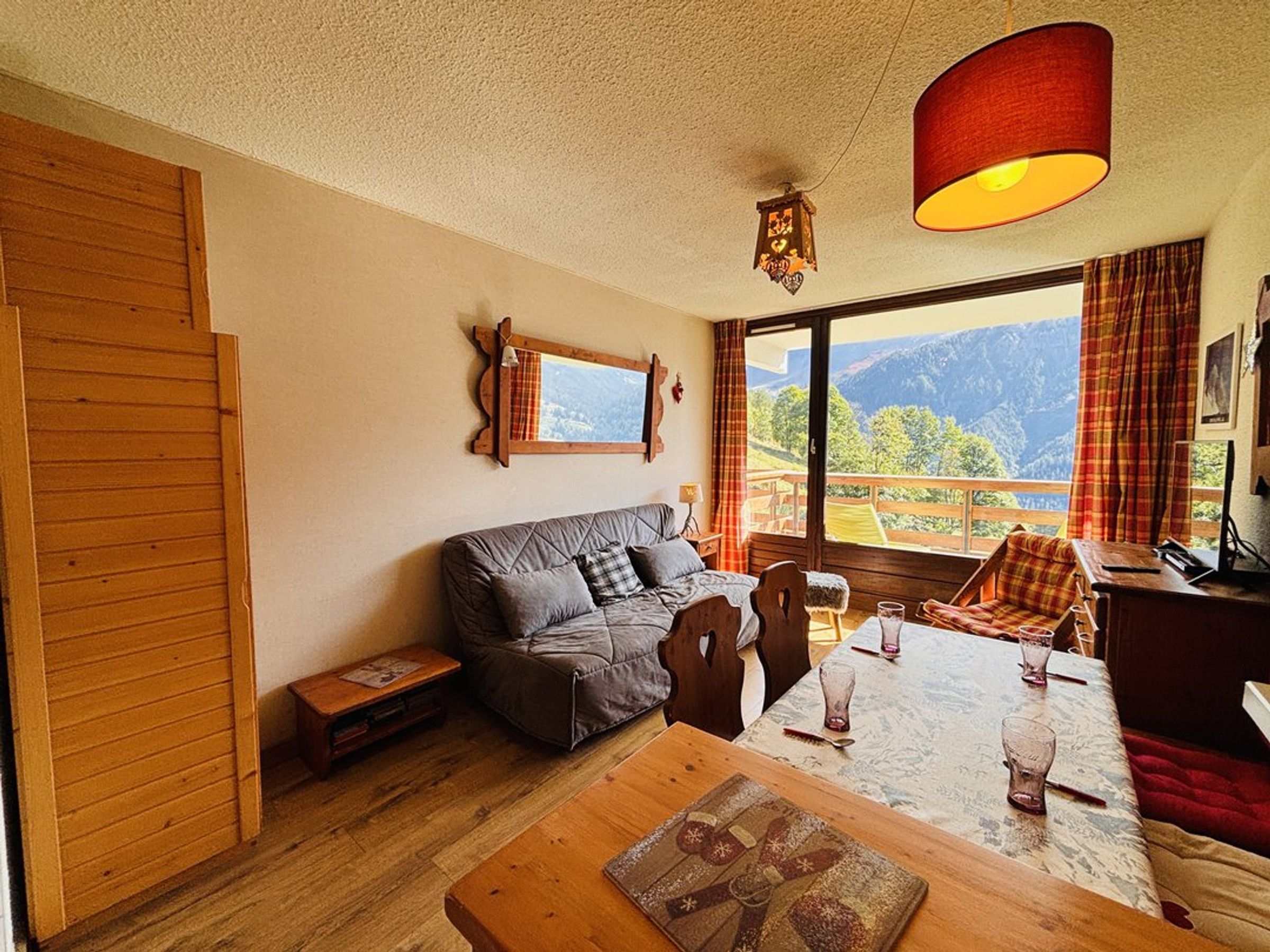 A cozy room with a wooden floor, a sofa, a dining table with chairs, and a large window offering a mountain view.