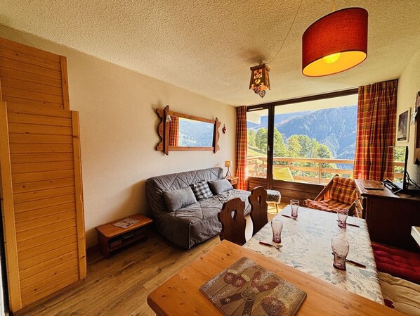 A cozy room with a wooden floor, a sofa, a dining table with chairs, and a large window offering a mountain view.
