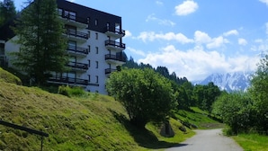 Un immeuble à plusieurs étages doté de balcons, entouré d’une végétation luxuriante et avec une chaîne de montagnes en toile de fond.