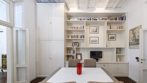 Una habitación con una mesa blanca, dos sillas, un florero rojo, una estantería, un cuadro enmarcado y un televisor.