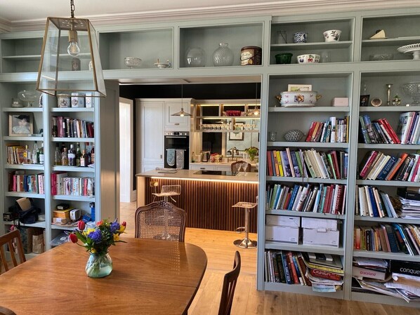 Uma área de refeições com uma mesa de madeira, cadeiras e um vaso de flores. Há estantes de livros em ambos os lados do quarto, repletas de livros e objetos decorativos.