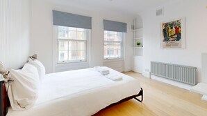 Una camera d'albergo con un letto grande, due finestre con tende, un termosifone, una ventola a soffitto e un quadro appeso al muro.