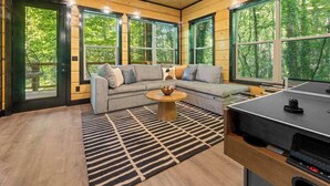 A cozy cabin interior with a sectional sofa, a round coffee table, and large windows offering a view of the forest. - Rustic Cabin Retreat・Sleeps 10・Indoor Pool・Hot Tub (Sevierville)
