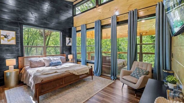 A modern bedroom with a large bed, a desk, and a chair. There are multiple windows with curtains, and a TV mounted on the wall. - Rustic Cabin Retreat・Sleeps 10・Indoor Pool・Hot Tub (Sevierville)