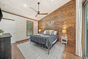 Un dormitorio con cama, ventilador de techo, pared de madera, ventana con persianas y mesita de noche con lámpara.