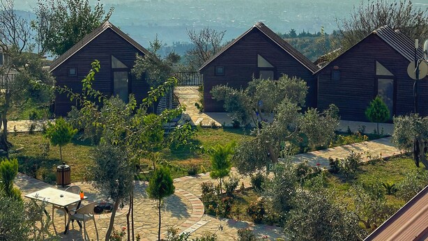A row of wooden cabins with a paved pathway in between, surrounded by greenery and a distant view of mountains. - LUNA HOME CLUB (Samandagi)
