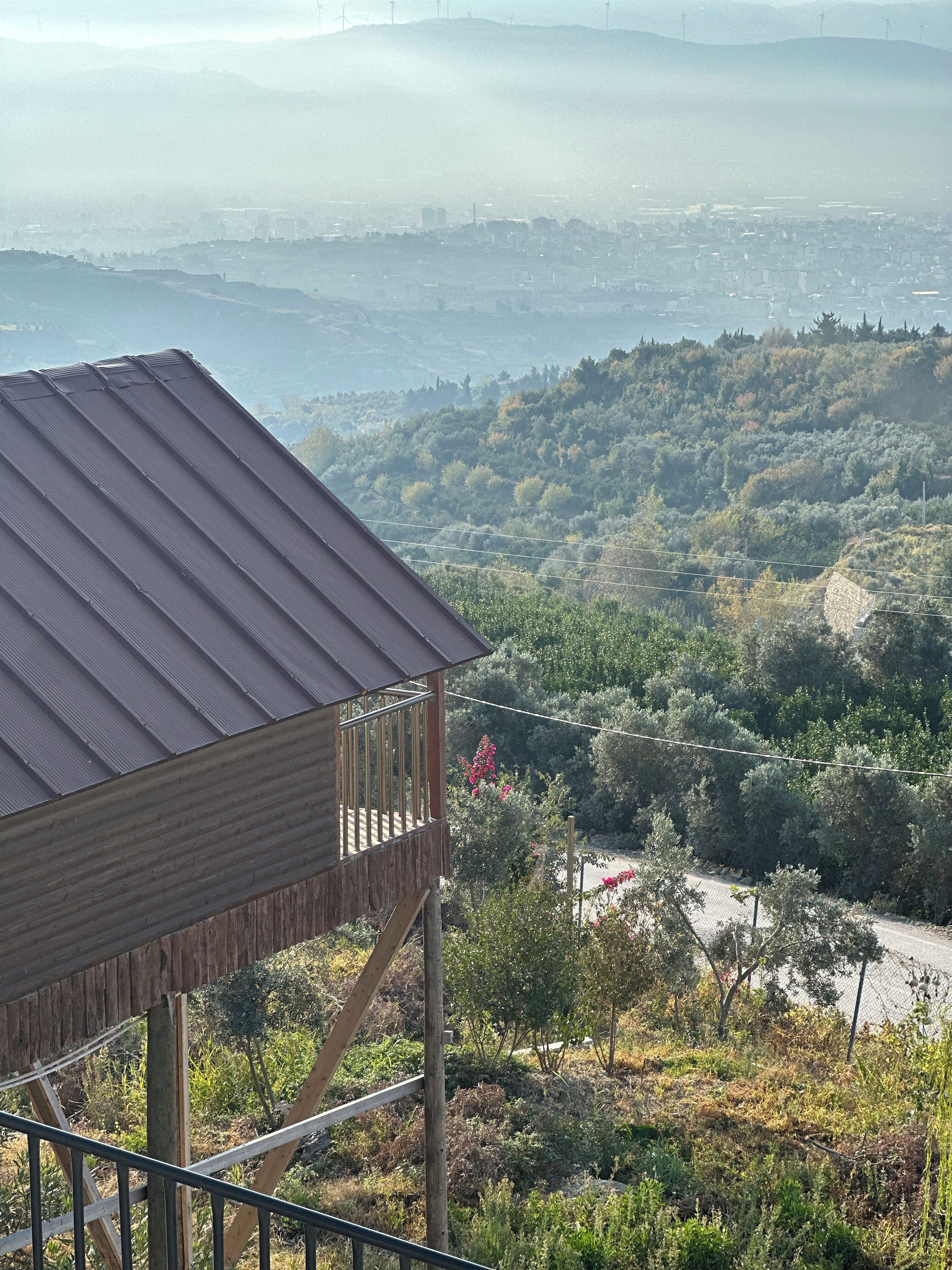 A wooden structure with a metal roof overlooking a hilly landscape with trees and a distant cityscape.