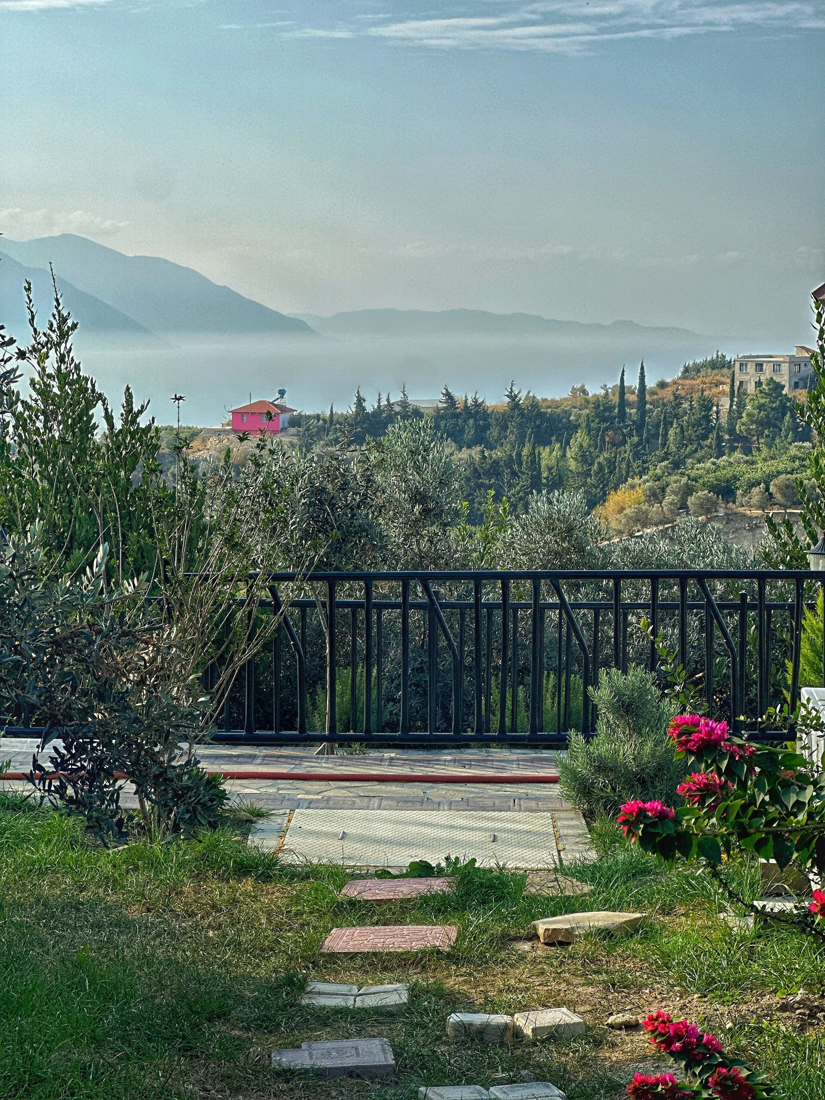 Ein Gartenweg führt zu einer Brücke mit Blick auf entfernte Gebäude und Berge.