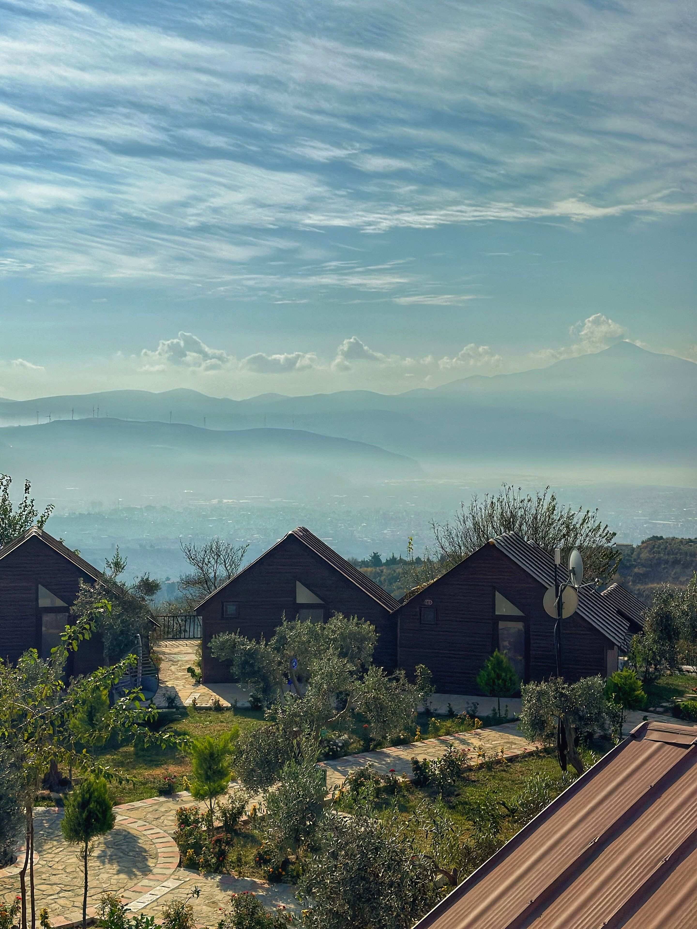Vista de tres cabañas de madera con un sendero de piedra que las conduce, rodeadas de vegetación y con vistas a una cadena montañosa distante.