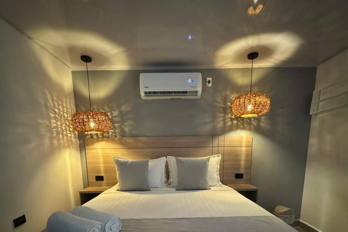 Quarto com uma cama grande, duas luminárias de cabeceira, um aparelho de ar condicionado e um ponto de luz fixado na parede.