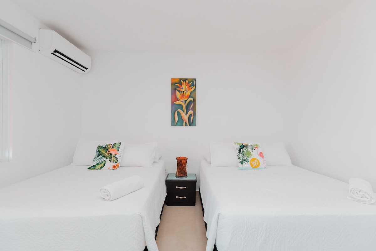 Une chambre d’hôtel avec deux lits, un climatiseur fixé au mur, une petite table avec un vase et un tableau au-dessus des lits.