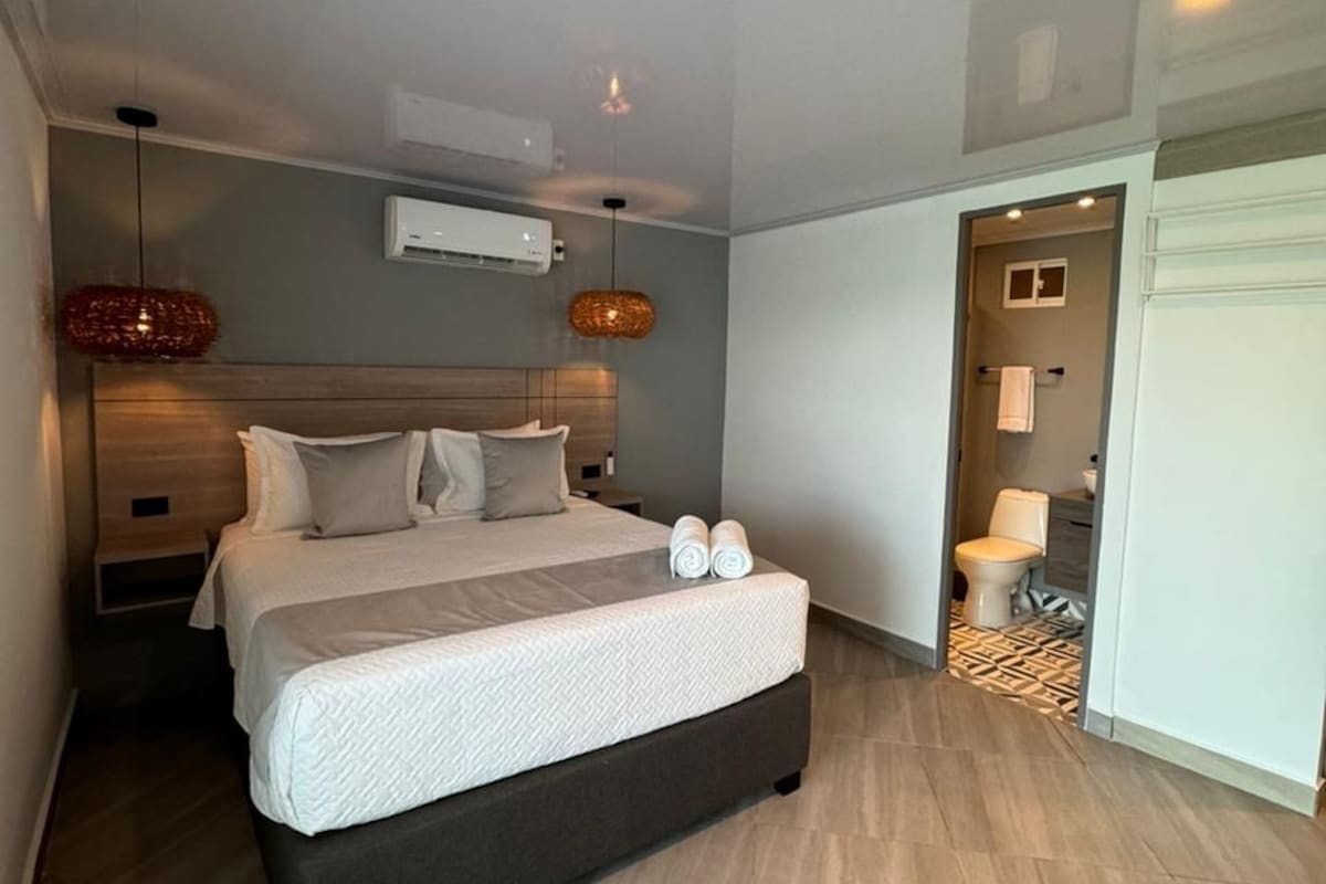 Une chambre moderne avec un grand lit, des tables de chevet, un appareil de climatisation et une salle de bain visible à travers une porte ouverte.