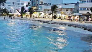 Un hotel frente a la playa con arena, palmeras y un paseo marítimo con tiendas y restaurantes.