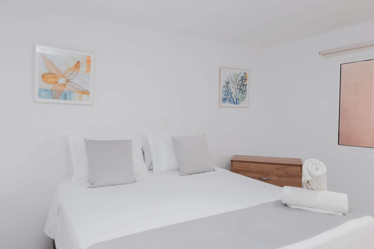 Quarto com cama, almofadas, mesa de cabeceira de madeira e duas obras de arte na parede.