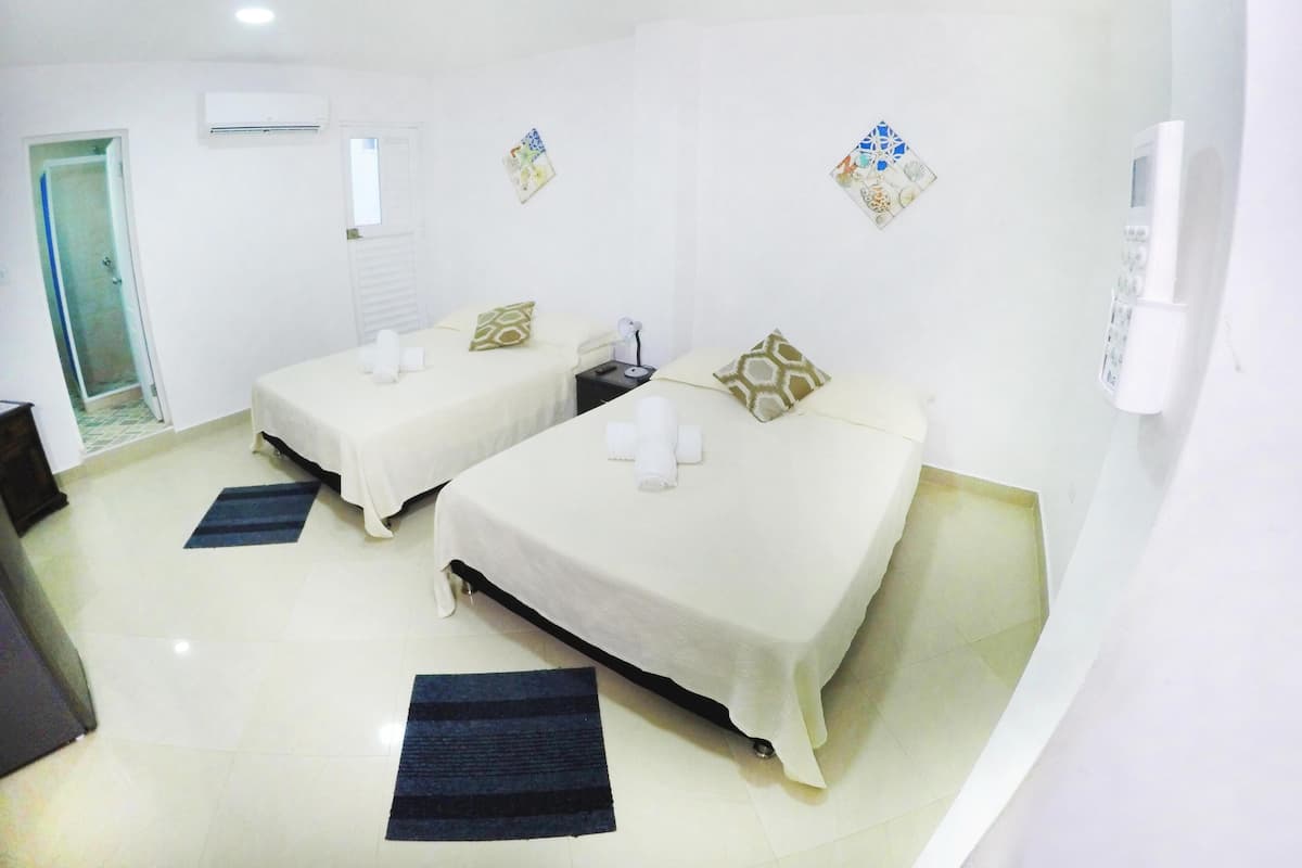 Quarto de hotel com duas camas, uma mesinha de cabeceira com abajur, telefone fixado na parede e uma porta que dá para o banheiro.