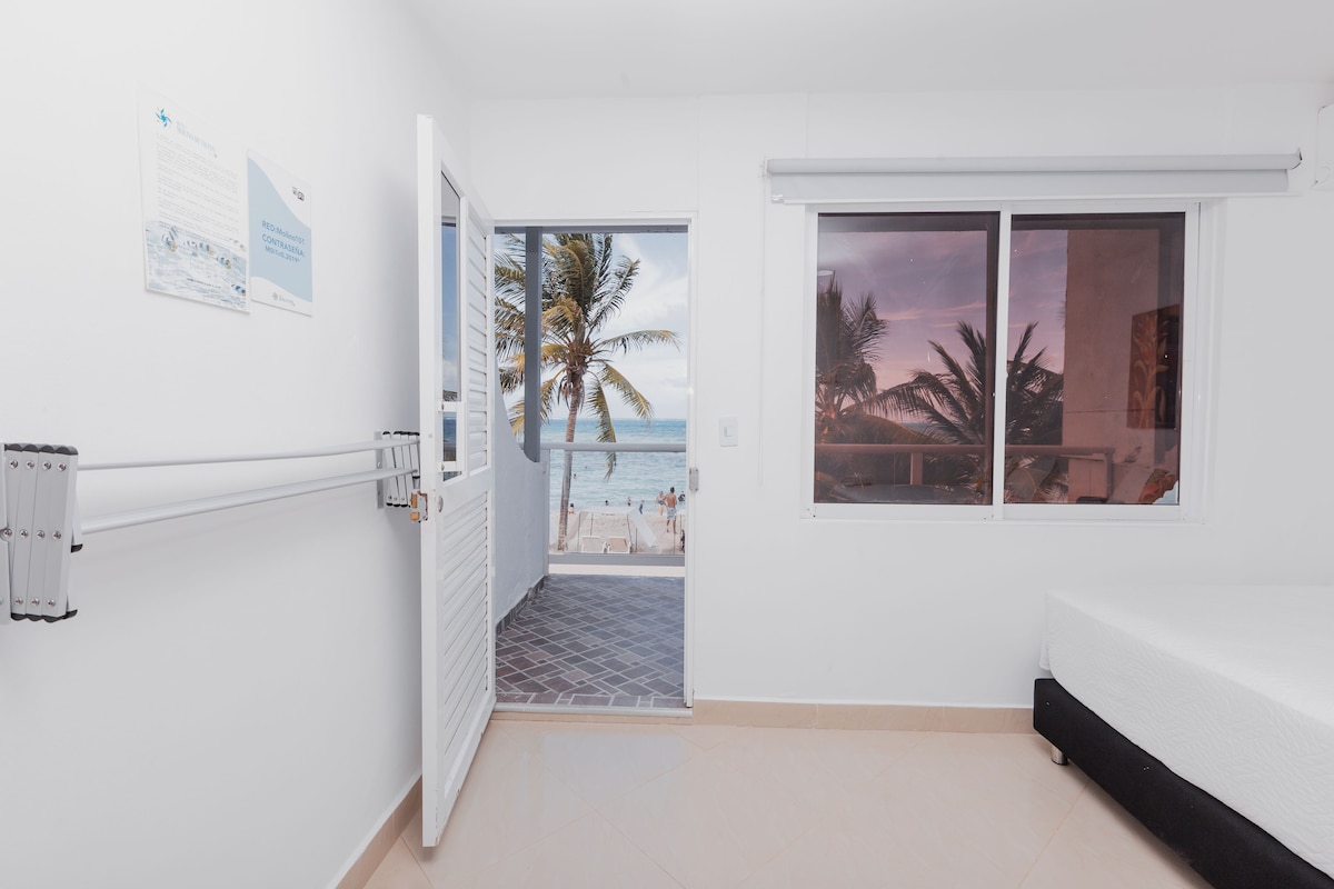 Une chambre avec un lit, une fenêtre donnant sur des palmiers et la plage, et une porte menant à un balcon.