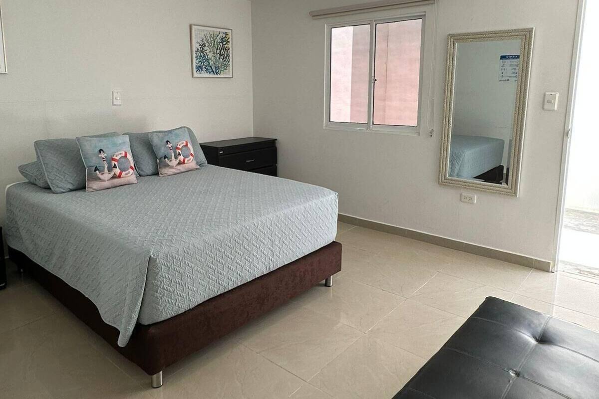Quarto com cama, mesa de cabeceira, cômoda, janela e espelho.