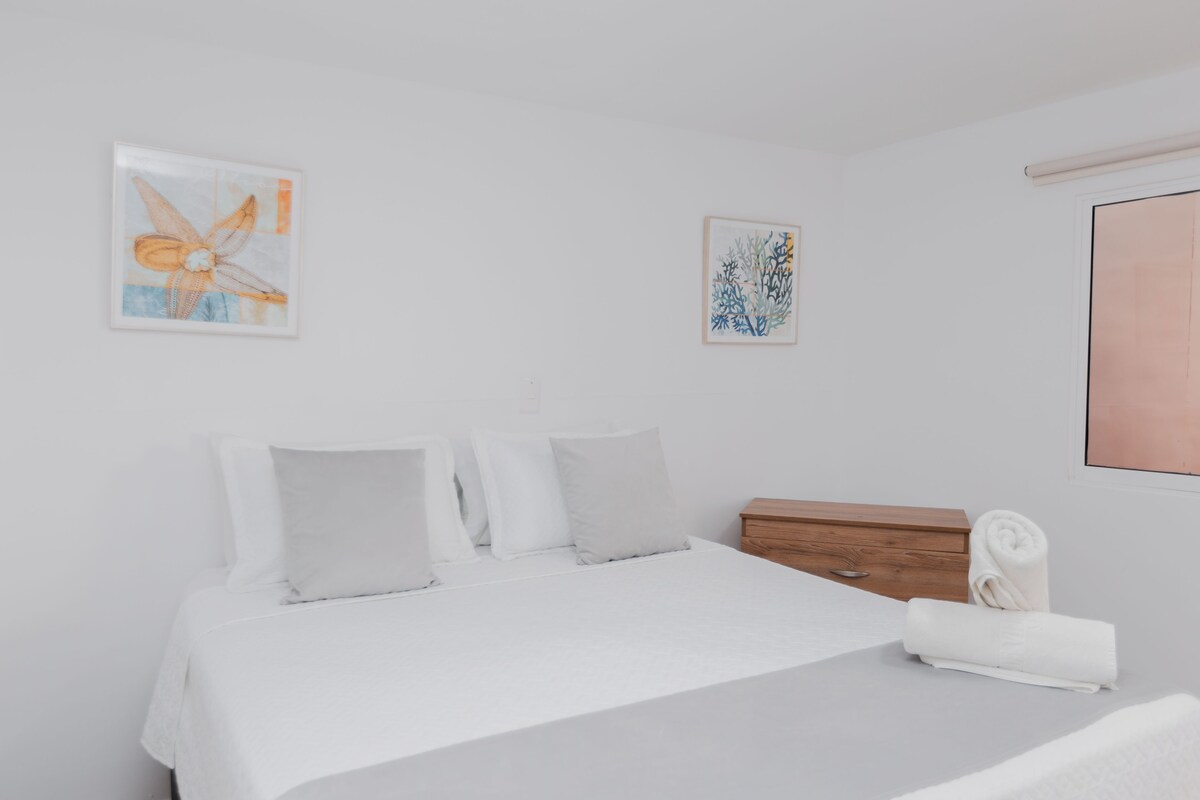 Une chambre à coucher comprenant un lit, des oreillers, une table de chevet en bois et deux œuvres d’art murales.