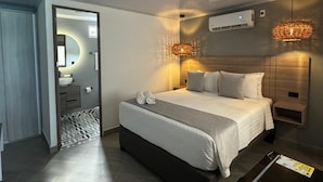 Una habitación de hotel moderna con una cama grande, mesitas de noche, un armario empotrado, un baño con espejo y lavabo, y un equipo de aire acondicionado.