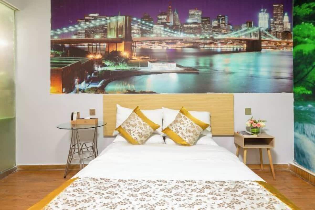Quarto de hotel com uma cama, um mural da cidade à noite, uma pequena mesa e uma mesa de cabeceira.